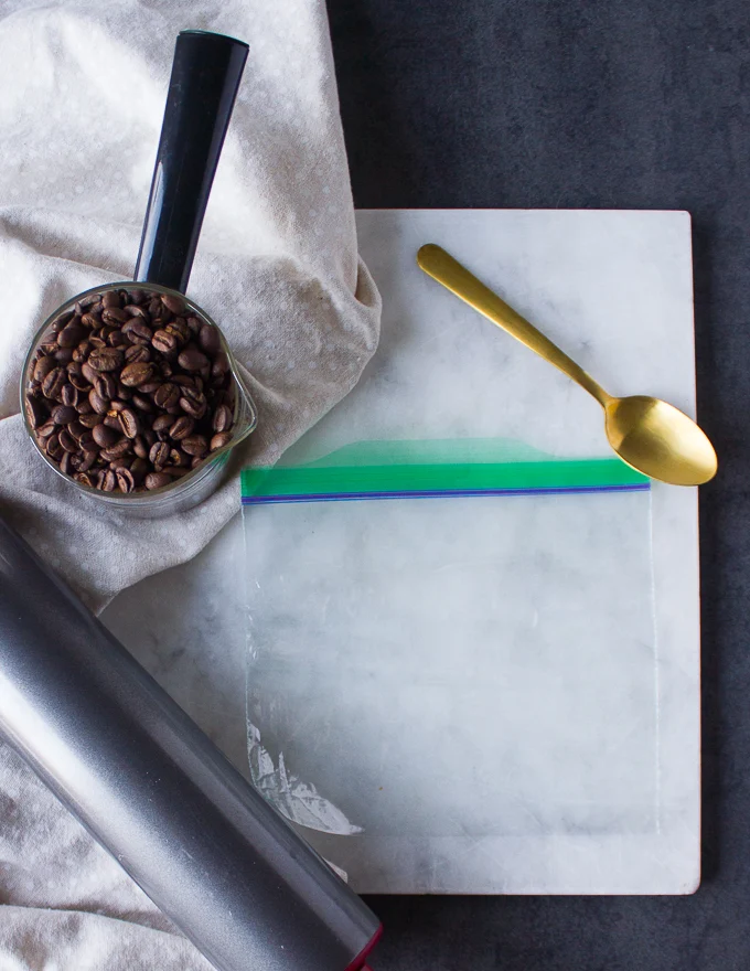 coffee beans in a cup and a ziploc over a white marble and a rolling pin to crush the coffee beans