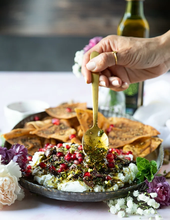 A hand spooning over the toppings over the spread out labneh over a serving plate with the pita chips