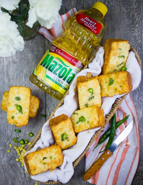 top view of a basket of cornbread muffins and a mazola corn oil bottle, a kitchen towel and some flowers in a glass