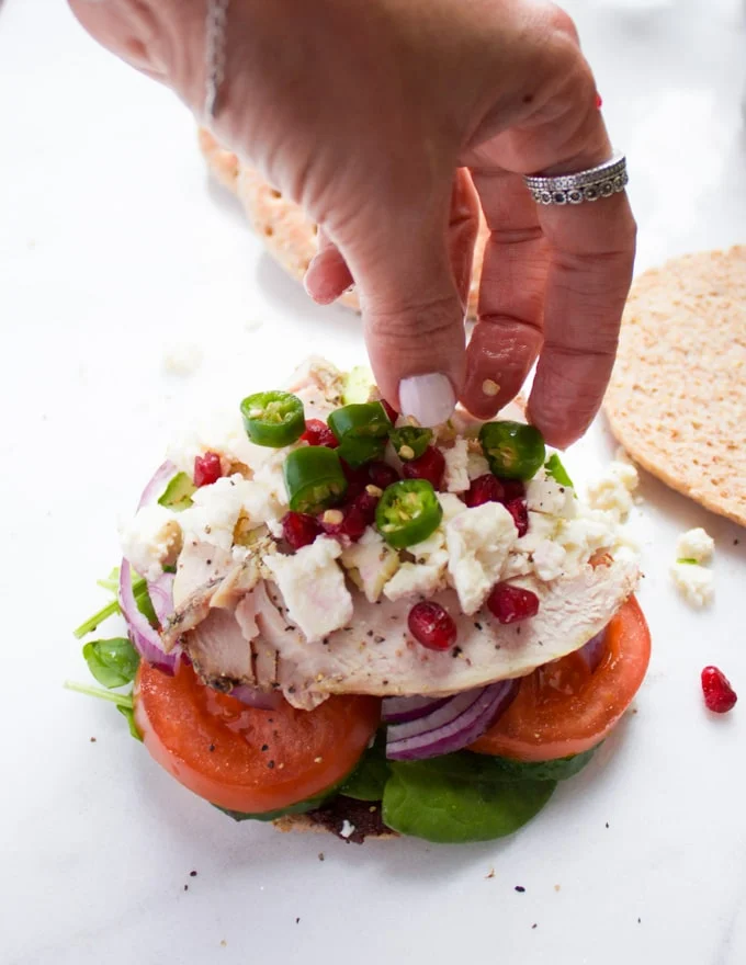 A hand finishing off the sandwich with a sprinkle of pomegranate arils, green chillies, feta cheese over turkey sandwich with cucumbers, tomatoes, red onions, spinach on whole wheat pita buns