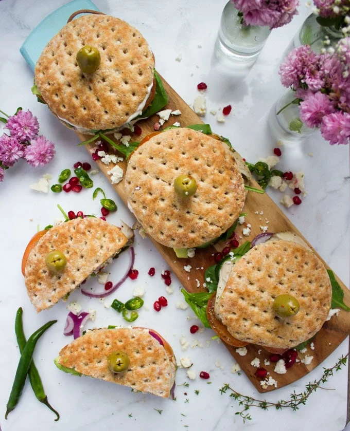 Top view of a white granite with a wooden board and turkey sandwiches over the wooden board. Another cut sandwich on the sandwich and flowers. Green chillies next to the cut up sandwich.
