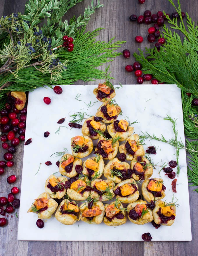 A tree shaped easy appetizer of mini pastries on a white marble board surrounded by fresh cranberries and green wreath leaves.