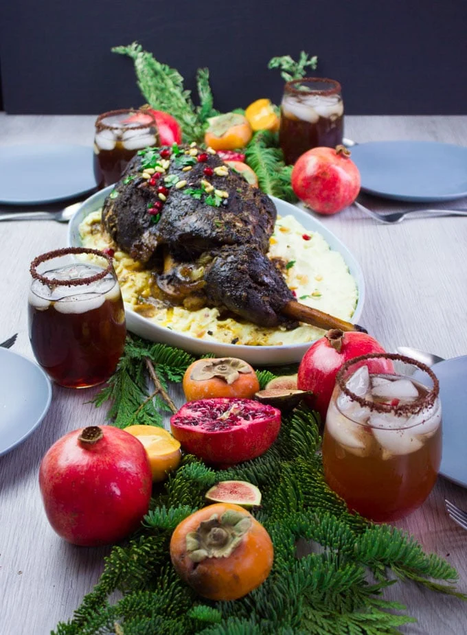 A few cups with drinks on a table and a few winter greens and fruits surrounding a big plate of a whole leg of lamb recipe served with mashed potatoes under.