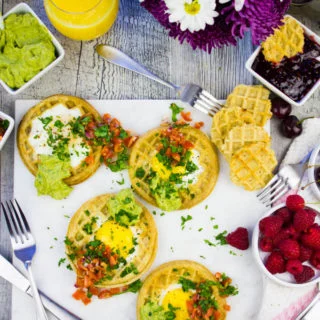 Finished breakfast table with four egg in a hole waffles, some flowers and berries, extra guacamole and jam on the side.