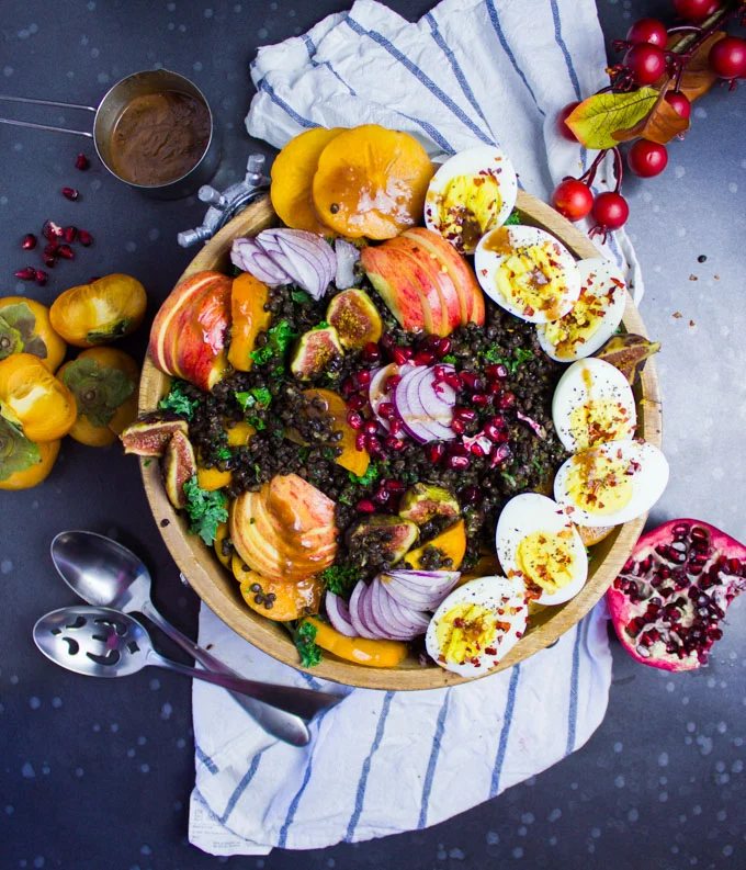 A top view of a kale salad bowl with fruits, hard boiled eggs, spicy lentils surrounded by fresh fruits, a kitchen towel and some spoons.