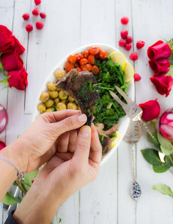 two hands sharing and holding a single lamb chop over a platter of lamb chops and roses, raspberries and drinks for a date night table set up