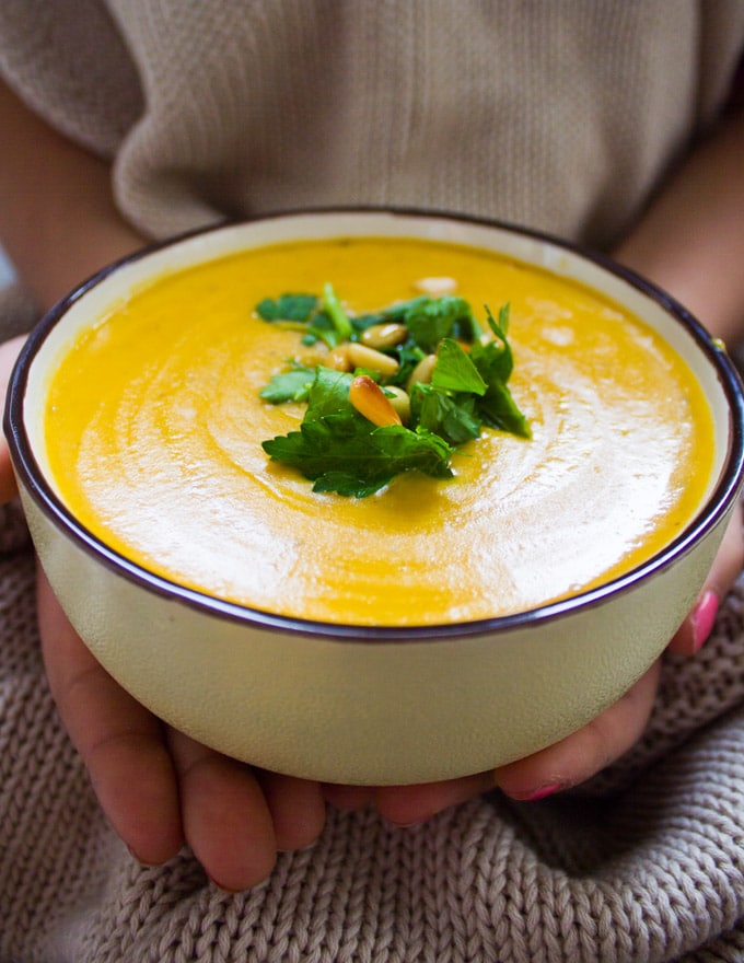 A hand holding a bowl of butternut squash soup recipe close to her showing the creamy soup and toppings.