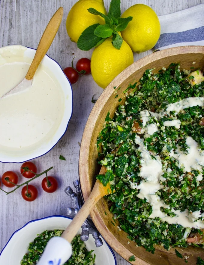 A half a bowl of tabouleh salad with lemons on a table and a bowl of tahini dressing