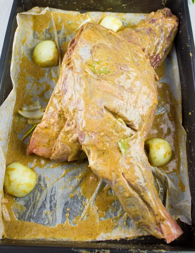 Fully marinated lamb shoulder on a sheet pan surrounded by whole onions and ready to roast