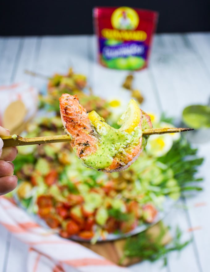 A hand holding the grilled salmon skewer right over a platter of salmon salad and drizzled with a dollop of dill dressing