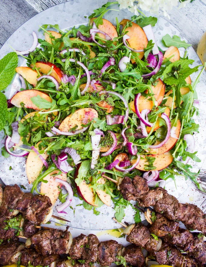 A plate of the peach salad surrounded by two grilled lamb skewers