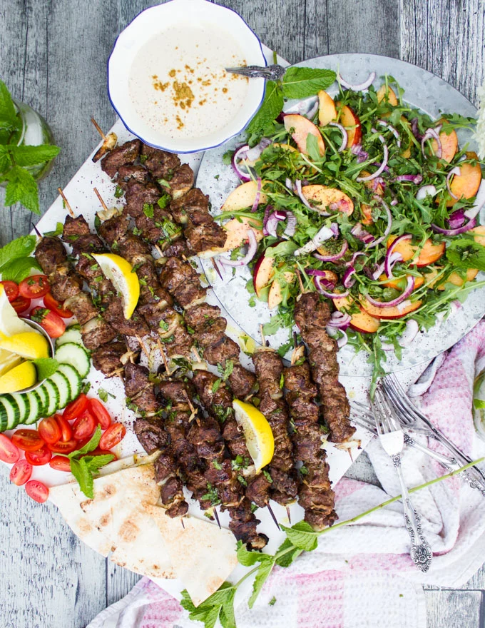 The BBQ party setup with grilled lamb skewers, peach salad, sliced cucumbers, tomatoes, tahini sauce and pita bread wedges