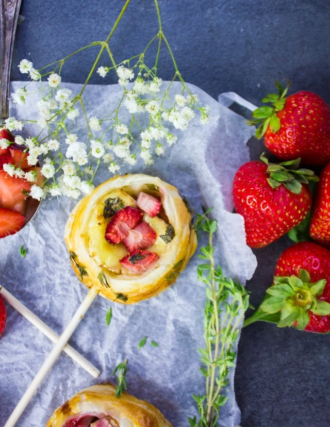A savory hand pie on a lollipop stick with strawberries surrounded by fresh strawberries, fresh thyme and white flowers