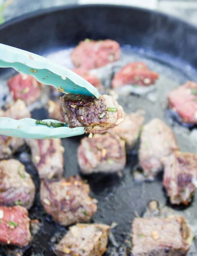 Steak Bites searing in a pan and a tong holding a piece of steak bite to show a close up of the searing