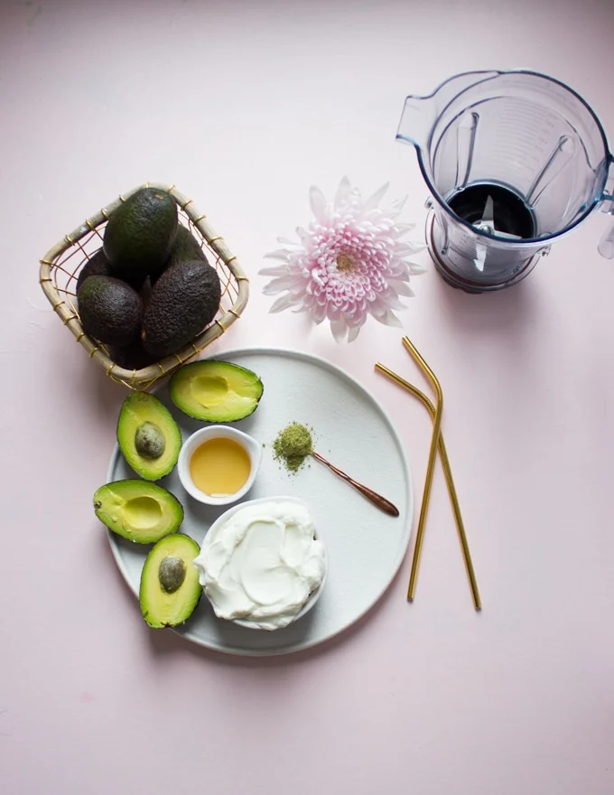 A plate with a cup fof yogurt, some avocados sliced in half, a little bit of honey, a teaspoon of matcha and a blender.f