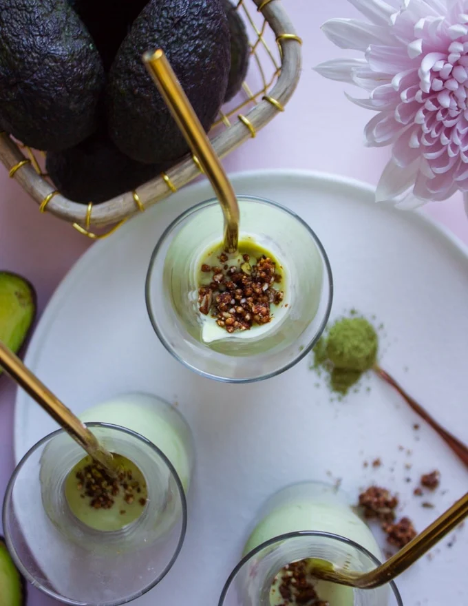 Three smoothie cups with straws and chocolate crumble on a white plate near a bowl of raw avocados