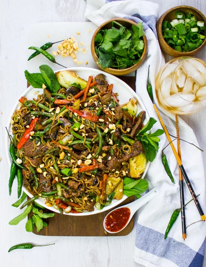A table set up with a plate of stir fry recipe, chilli sauce on the same, cold drinks, extra cilantro and mint for garnish, chopsticks