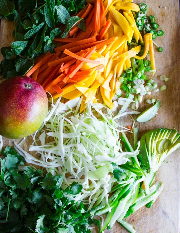 a wooden board with all the salad ingredients chopped: cabbage, mango , carrots, onions, mint leaves, cilantro leabes