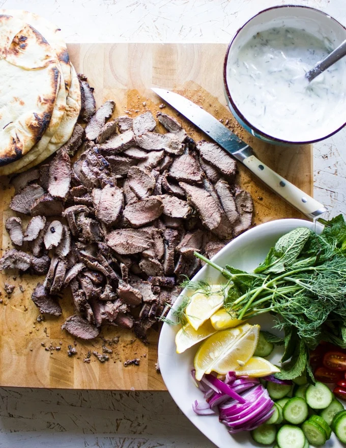 A board with all the gyro recipe ingredients: Sliced lamb sirloin, charred pita bread, Tzatziki sauce, a plate of sliced cucumbers, tomatoes, fresh herbs, sliced onions and lemon slices