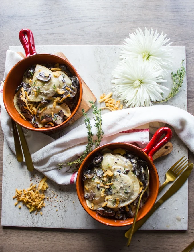 two bowls of pierogies alfredo recipe served with spoons and forks , surrpunded by a tea towel, white flowers and fresh thyme leaves