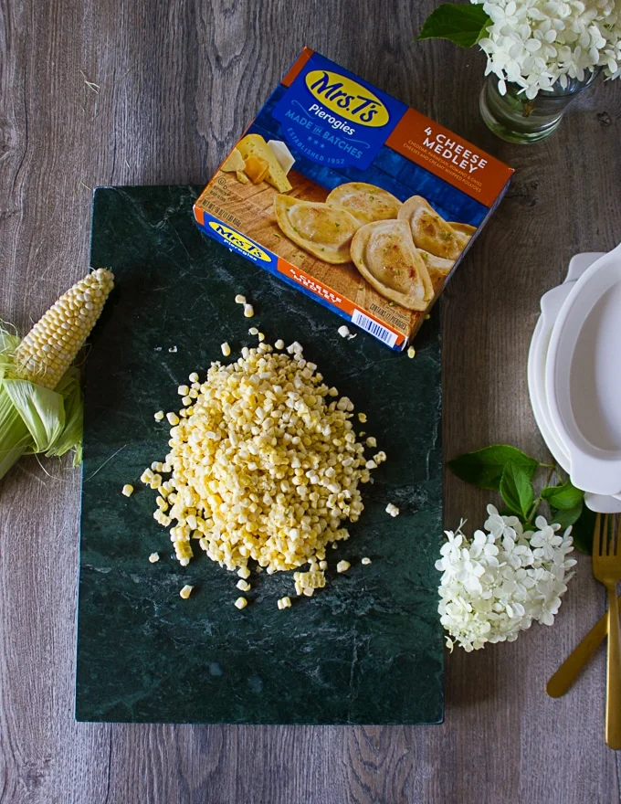 Ingredients for corn casseole over a marble board: the pierogies box, corn kernels, cream, ramekins and garlic