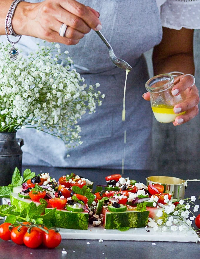 A hand holding a jar of Greek salad dressing and drizzling the dresisng with a spoon over the watermelon salad