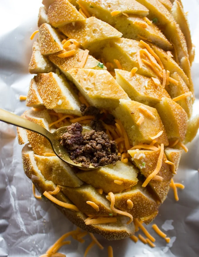 A spoon adding in the cooked lamb crumbles over the cheese in the pull apart bread