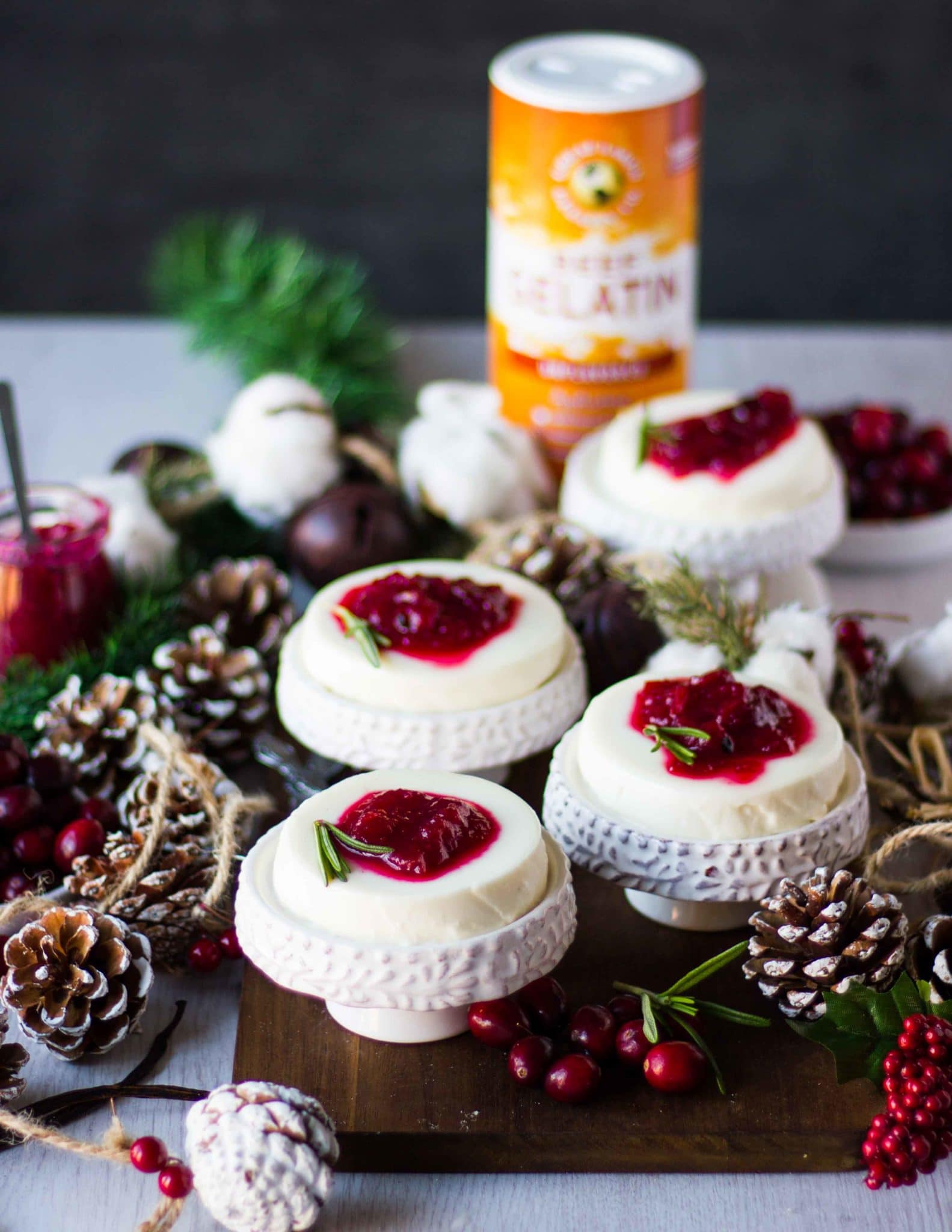 individual servings of four panna cotta recipe on serving plates finished with cranberry sauce and surrounded by fresh cranberries and wooden cones