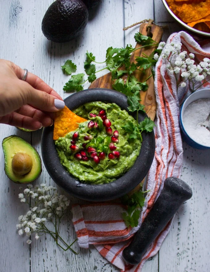 A hand holding a chip and dipping it into a guacamole dip in a bowl surrounded by a tea towel and hass avocados