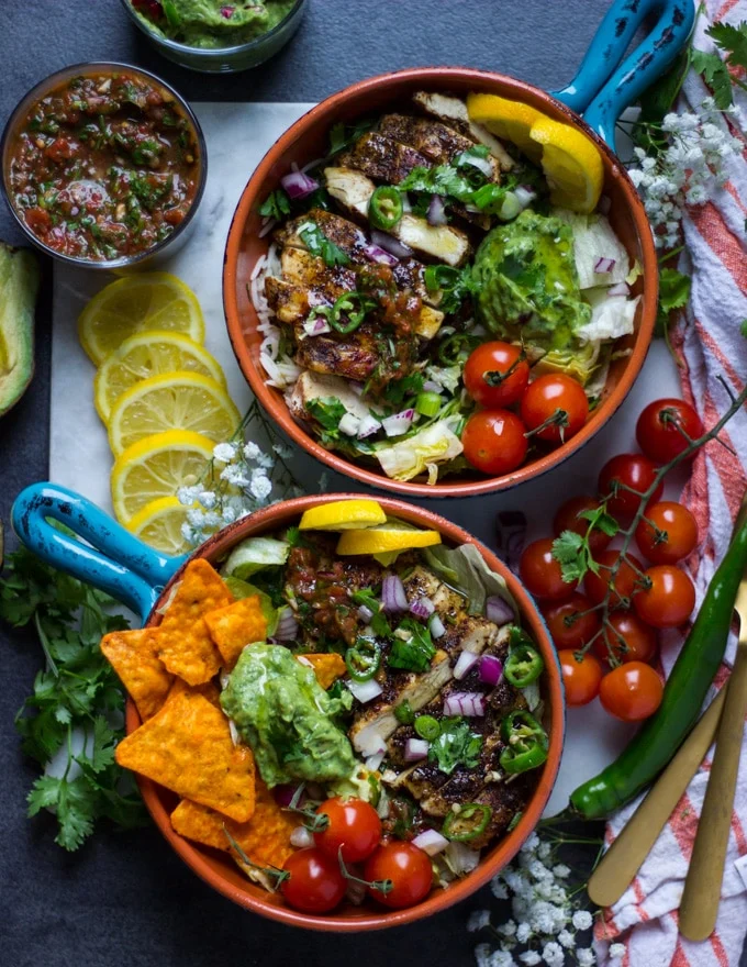 2 ready fajita bowls assembled with the toppings like guacamole, salsa, onion, tomatoes, jalapeno, surrunded by lemon slices, cilantro