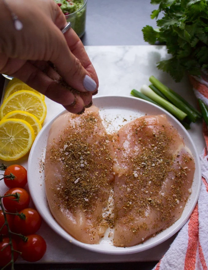 A hand sprinkling some fajita spice over the raw chicken breast to flavor them