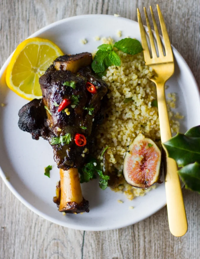 A serving plate with a piece of cooked lamb shoulder with Moroccan spice in a sweet and spice sauce surrounded by some couscous and fresh figs and a slice of lemon