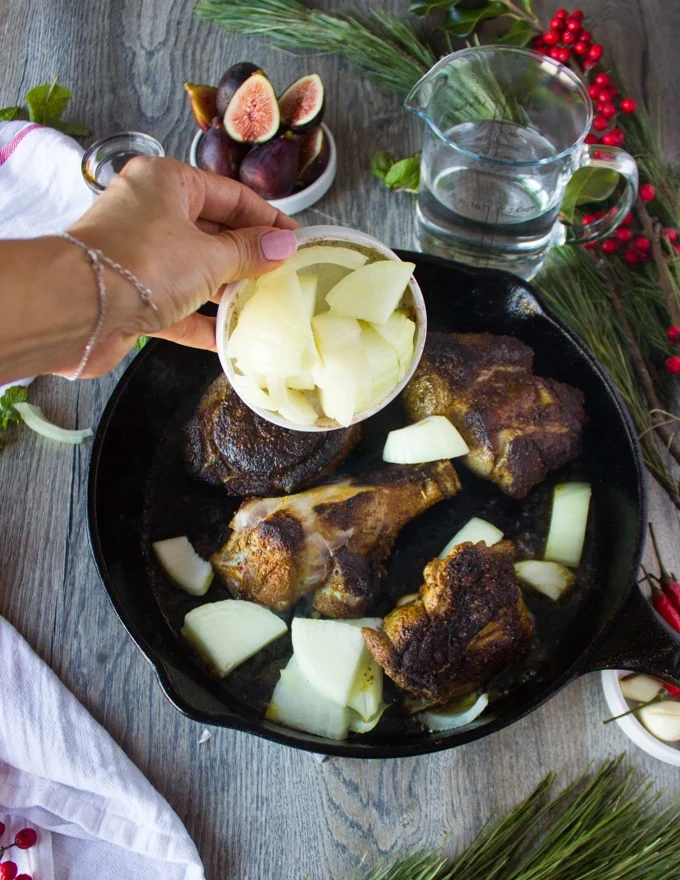 A hand pouring chopped onions over the seared lamb in a skillet