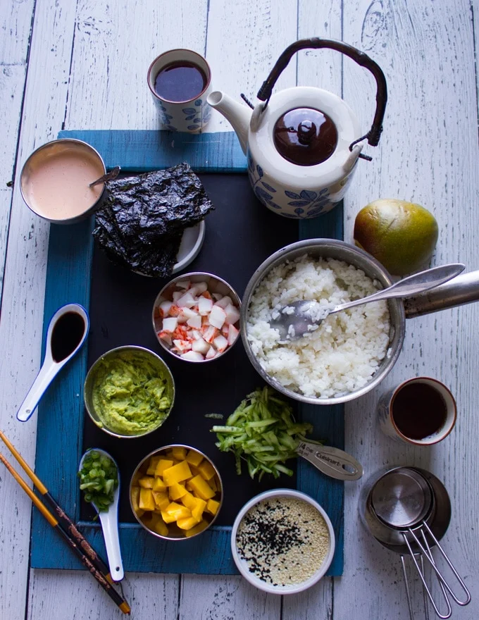 Ingredients for the crab sushi and sushi stacks, a pot of rice, seaweed sheets, spicy mayo, soy sauce, crab meat, mashed avocado, mango, scallions, shredded cucumbers, sesame seeds, dry measuring cups