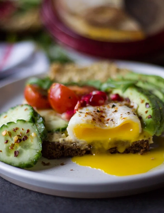 A cut through the breakfast toast showing the runny yolks from the poached egg over the hummus toast on a white plate surrounded by avocados, cucumbers and tomatoes