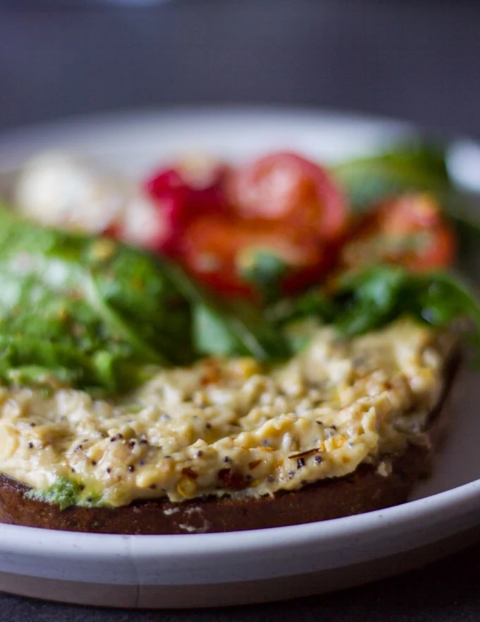 A loaded hummus toast or breakfast toast with a thick spread of hummus, some arugula, tomatoes, cucumbers, avocados and poached egg