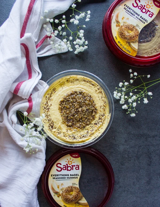An opened up tub of hummus showing the everything bagel seasoning swieled at the top of the hummus, surrounded by a tea towel