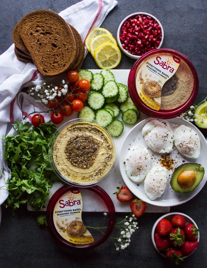 Ingredients for the hummus toast or breakfast toast on a white marble. Ingredients are a plate of poached eggs, a tub of hummus, sliced avocados, cucumbers, arugula, cucumbers, tomatoes, rye bread and some strawberries in a bowl , and lemon slices