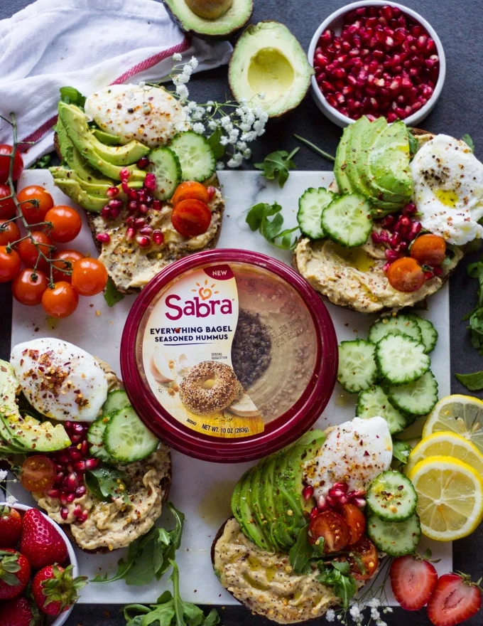 A marble plater with the fully assembled hummus toasts or breakfast toasts, each toast loaded with hummus, poached eggs, avocados, cucumbers, greens, tomatoes and sprinkles of pomegranate arils. A bowl of strawberries on the side, lemon slices and a tub of hummus in the center of the marble board.