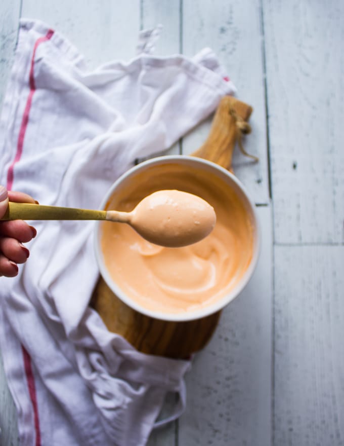 A hand holding a spoonful of spicy mayo over a bowl of spicy mayo recipe surrounded by a tea towel on a white board