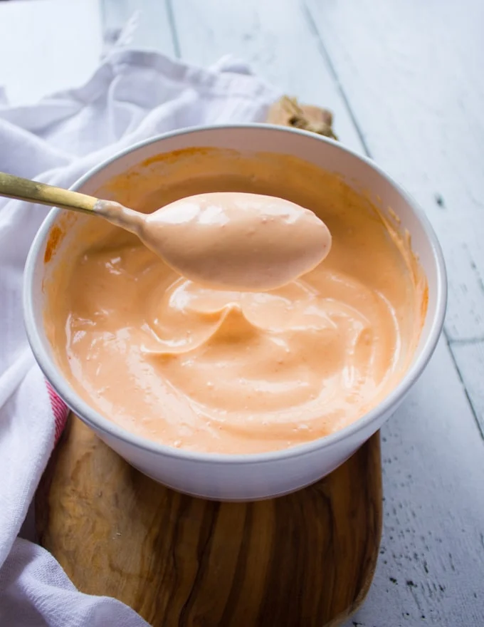 A spoon lifting up some spicy mayo sauce showing the thick drips of sauce over a spicy mayo bowl
