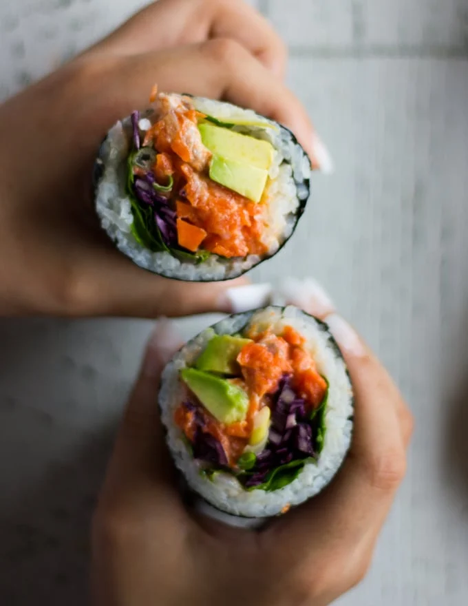 two hands holding off two halves of the sushi burrito pointing up and showing the contrast in colors of the fillings and the sushi rice and all the layers