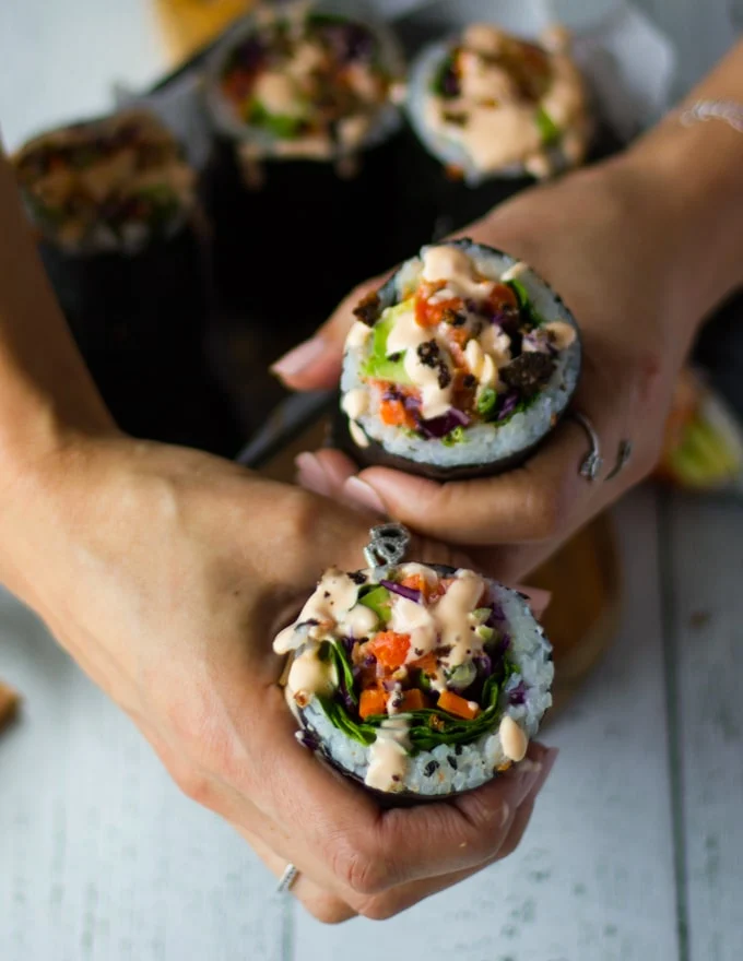 two hands holding the sushi burrito finished off with a drizzle of spicy mayo and the sprinkles of crispy salmon skin on top