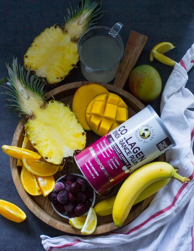 A wooden tray with all the tropical fruits used in the tropical smoothie such as half a pineapple, mangoes, cherries, banana, orange segments and coconut water and a box of collagen to boost the smoothie