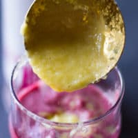A ladle spooning in the mango banana or pineapple banana smoothie layer of the cherry smoothie layer in the smoothie cup