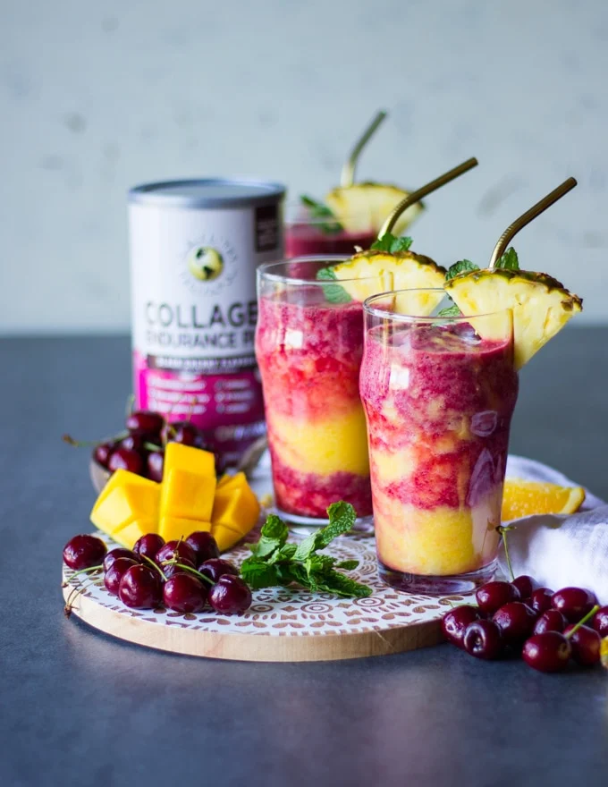 two thick tropical smoothie cups on a white wooden board surrounded by fresh cherries and mangoes with two straws and sliced pineapple for garnish