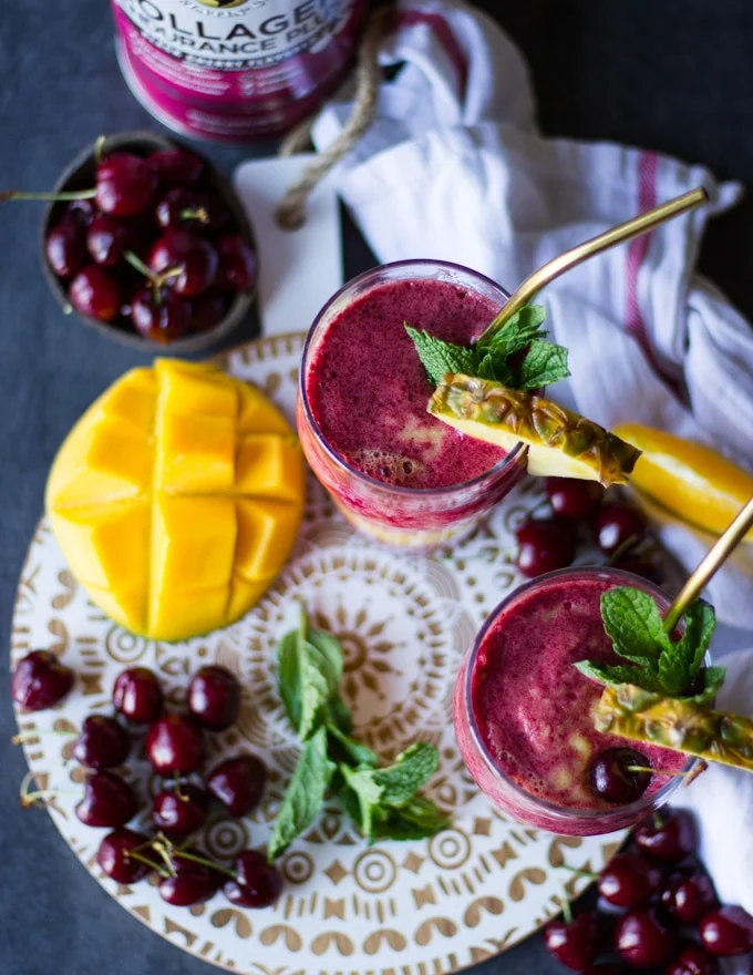 Top view of two smoothie cups showing the thickness and color blends of the cherry smoothie and mango banana smoothie surrounded by fresh mint and more tropical fruits and cherries