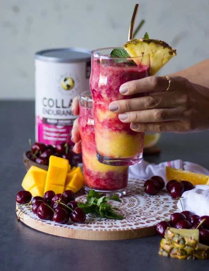A hand holding up a cup of tropical smoothie highlighting the layers of smoothies. Some fruit surrounded the table like fresh cherries and mangos