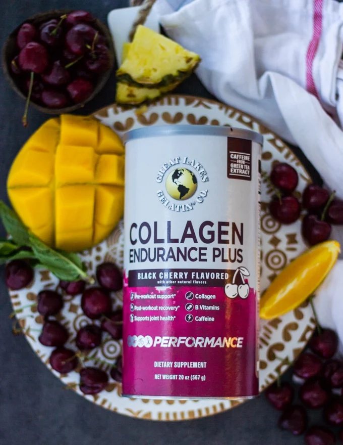 A box of GLG Collagen Endurance surrounded by fresh cherries, mangoes, pineapple slices and an orange segment on a white wooden board