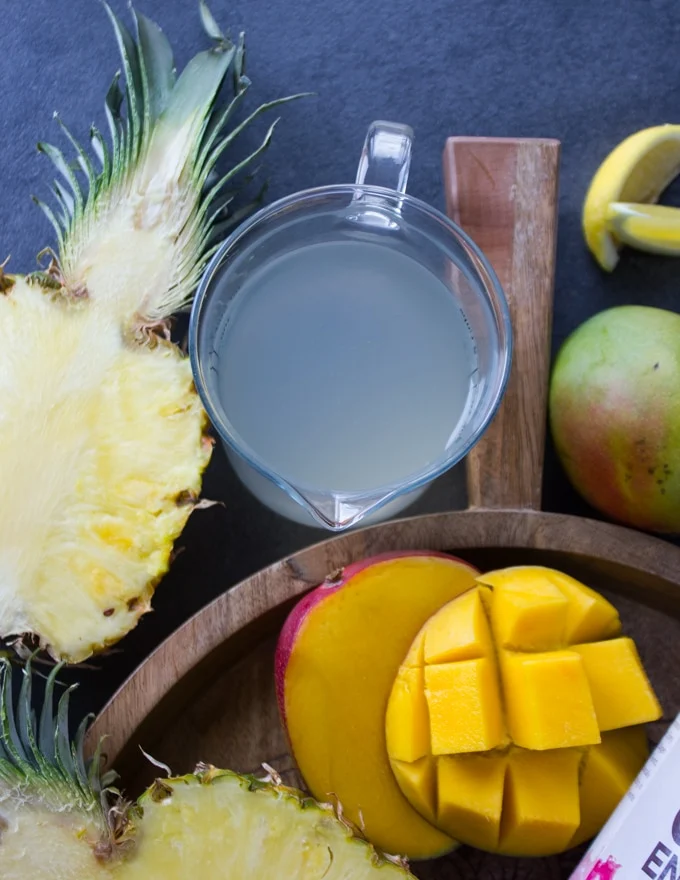 A cup with coconut water surrounded by tropical fruits like half a sliced pineapple and mangos.